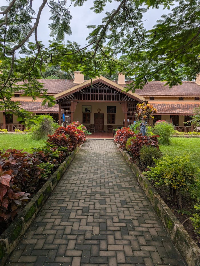Colonial-style bungalow surrounded by vibrant gardens, featuring a paved walkway under lush trees.